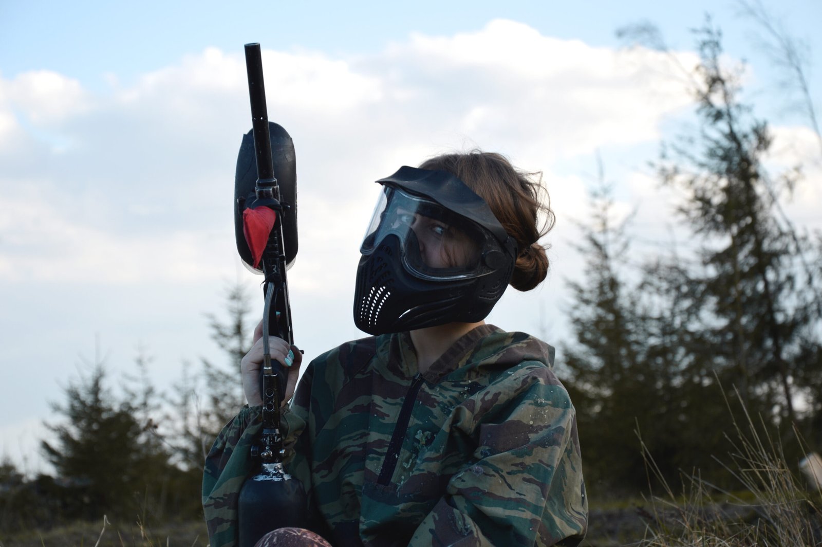 Paintball sport player girl in protective camouflage uniform and mask with marker gun outdoors